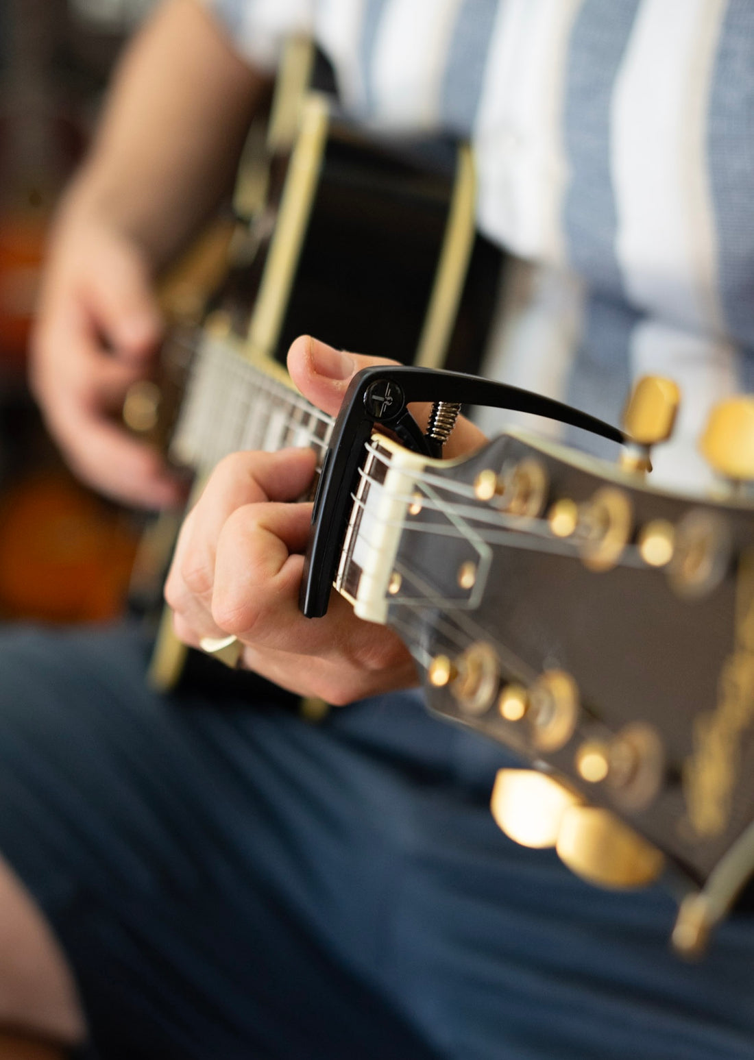 Een capo op een gitaar die gespeeld wordt door iemand met een streepjes blouse aan.